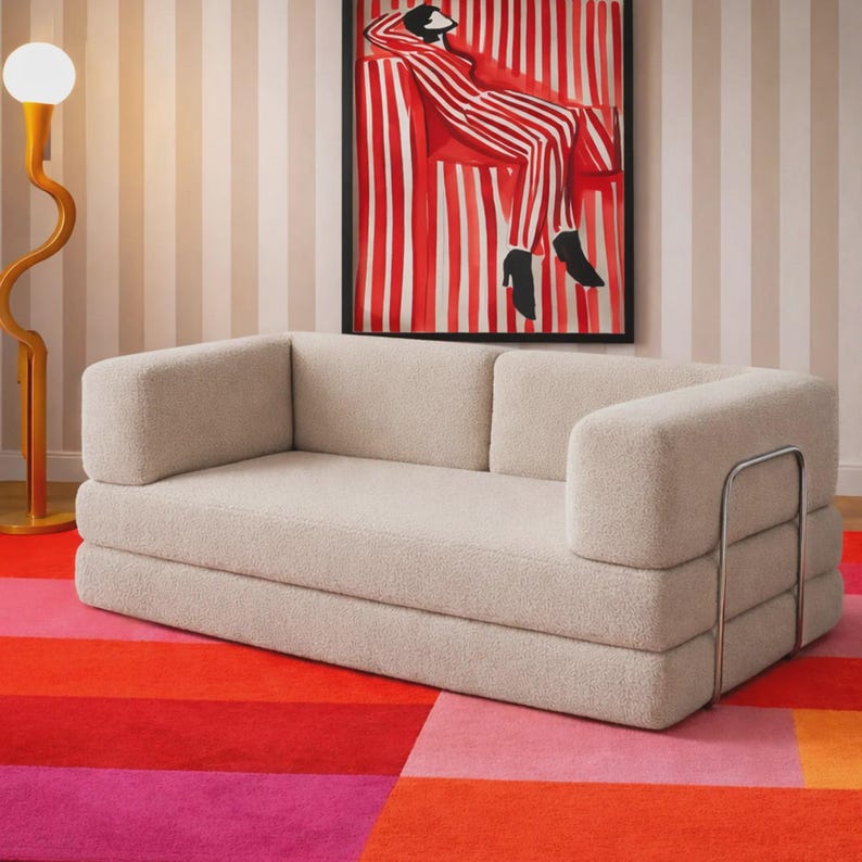 May include: A cream-colored, textured fabric sofa with a low-profile, rectangular design. The sofa features a metallic silver frame on the right side and a thick, cushioned seat and backrest. It is positioned in a room with a striped wall and rug.