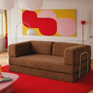 May include: A brown, textured, fuzzy fabric sofa with a low back and thick, rectangular armrests. The sofa features a silver-toned metal frame structure on the sides and is positioned on a bright red circular rug in a room with white walls.