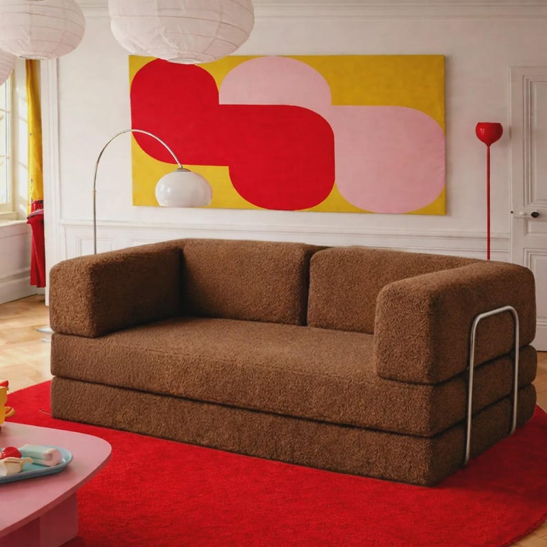 May include: A brown, textured, fuzzy fabric sofa with a low back and thick, rectangular armrests. The sofa features a silver-toned metal frame structure on the sides and is positioned on a bright red circular rug in a room with white walls.