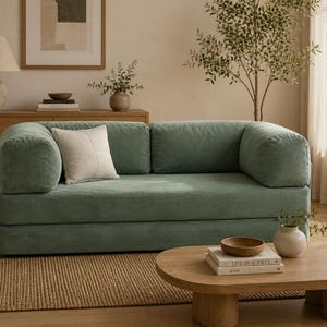 May include: A sage green corduroy sofa with rounded armrests and a single square off-white throw pillow. The sofa features a low-profile design with two back cushions and a continuous seat cushion, upholstered in a textured ribbed fabric.
