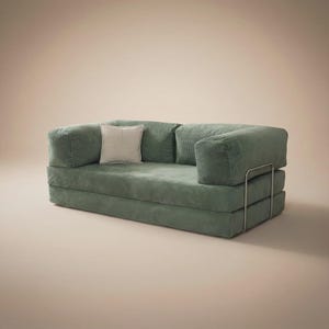 May include: A sage green corduroy sofa bed featuring a low-profile design with thick, cushioned armrests and a backrest. The piece includes a single square off-white throw pillow and is supported by a thin, silver-toned metal frame structure.