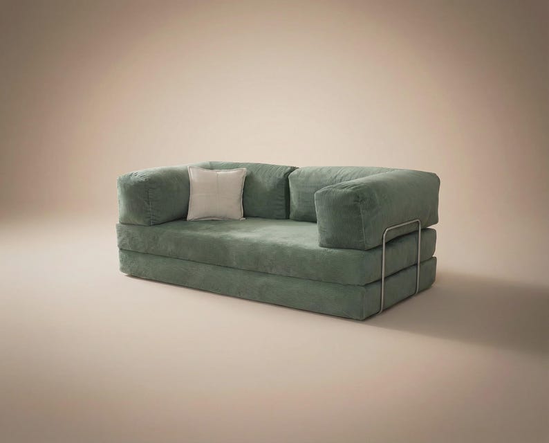 May include: A sage green corduroy sofa bed featuring a low-profile design with thick, cushioned armrests and a backrest. The piece includes a single square off-white throw pillow and is supported by a thin, silver-toned metal frame structure.