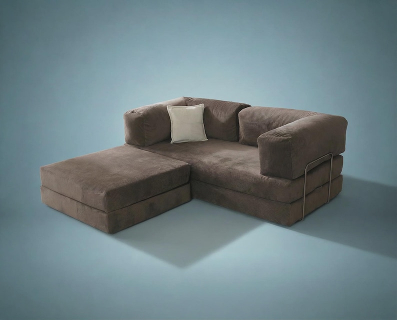 May include: A brown modular sofa with a low-profile design, featuring a chaise lounge section, thick cushioned armrests, and a single square off-white throw pillow. The sofa is supported by a thin, visible metal frame structure on the side.