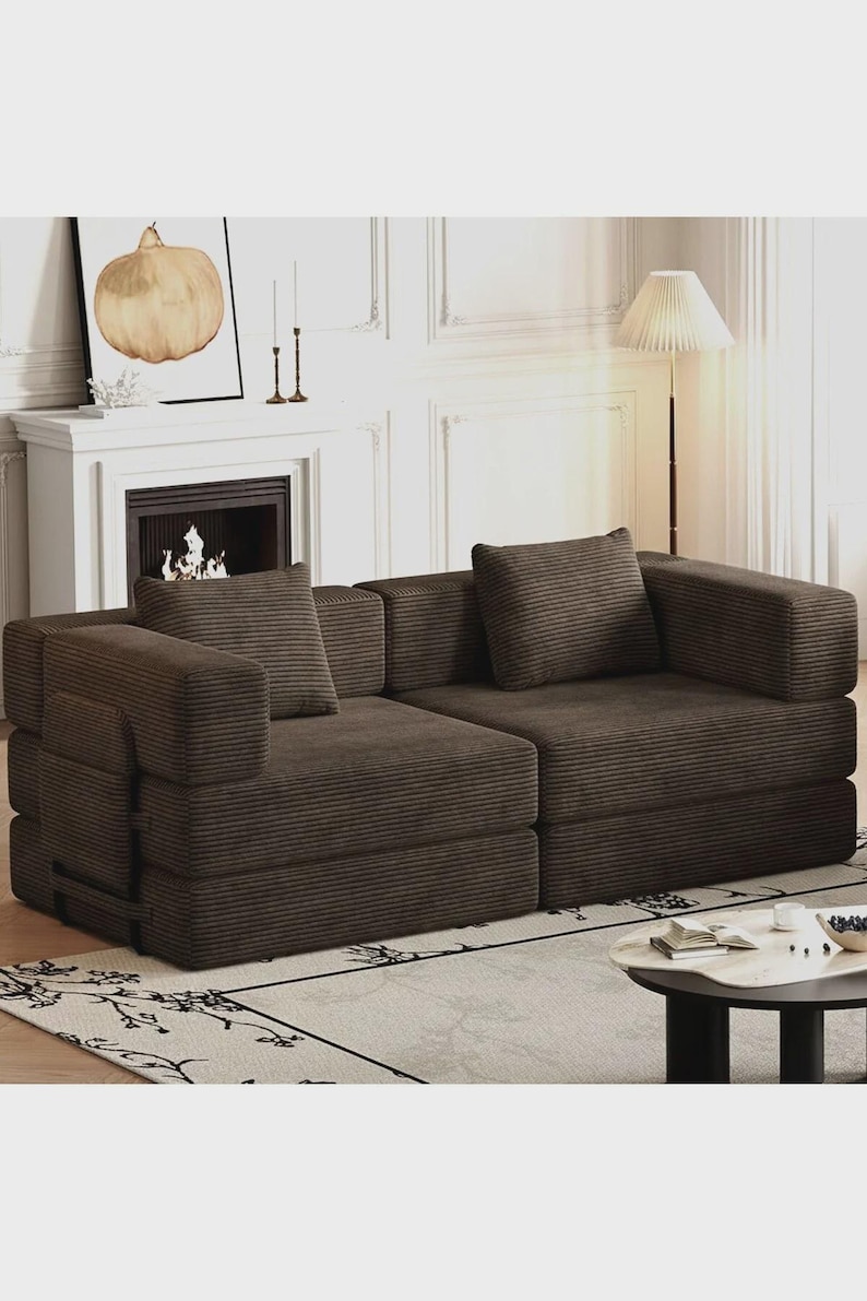 May include: A dark brown corduroy sofa featuring a low-profile design with two distinct seating sections, thick square armrests, and two matching square throw pillows. The fabric has a prominent vertical ribbed texture throughout the entire piece.