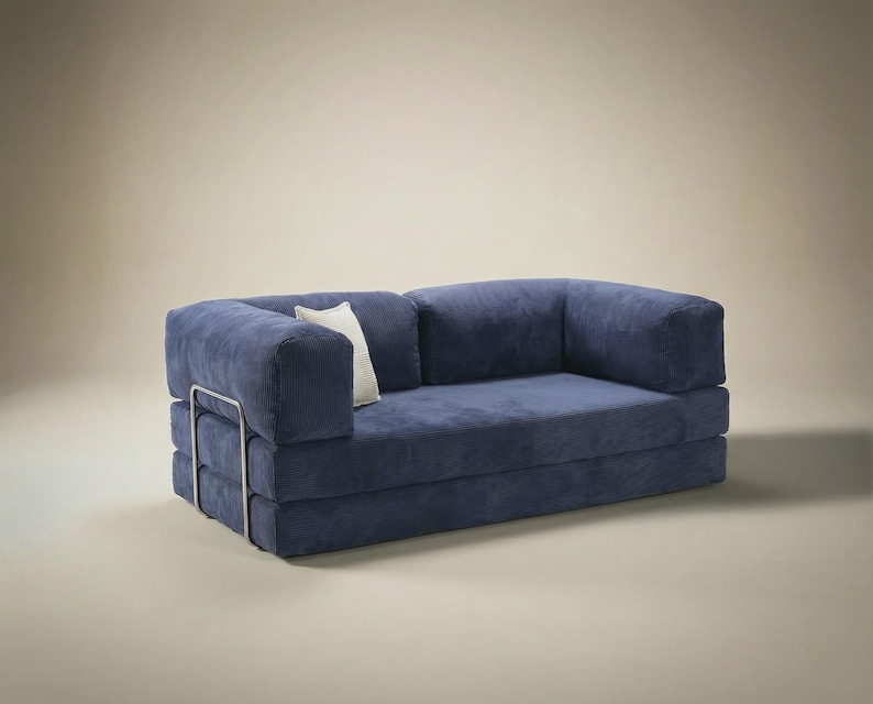 May include: A navy blue corduroy sofa featuring a low-profile design with thick, blocky cushions and a metallic silver frame visible on the side. A single small, off-white rectangular throw pillow is tucked against the left armrest.