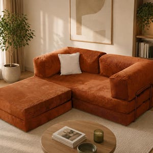 May include: A modular sofa upholstered in rust-colored corduroy fabric, featuring a low-profile design with a matching ottoman section, thick square armrests, and a single white square throw pillow placed against the back cushion.