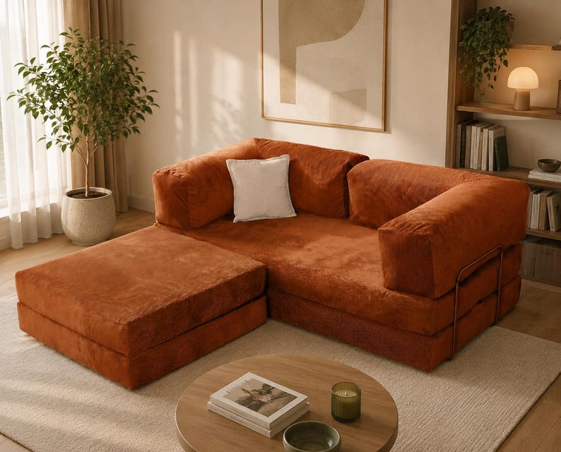 May include: A modular sofa upholstered in rust-colored corduroy fabric, featuring a low-profile design with a matching ottoman section, thick square armrests, and a single white square throw pillow placed against the back cushion.