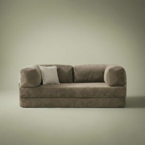 May include: A low-profile, taupe-colored sofa featuring a soft, textured fabric finish, rounded armrests, and two back cushions. A single, smaller square throw pillow in a lighter, off-white shade is positioned against the left back cushion.