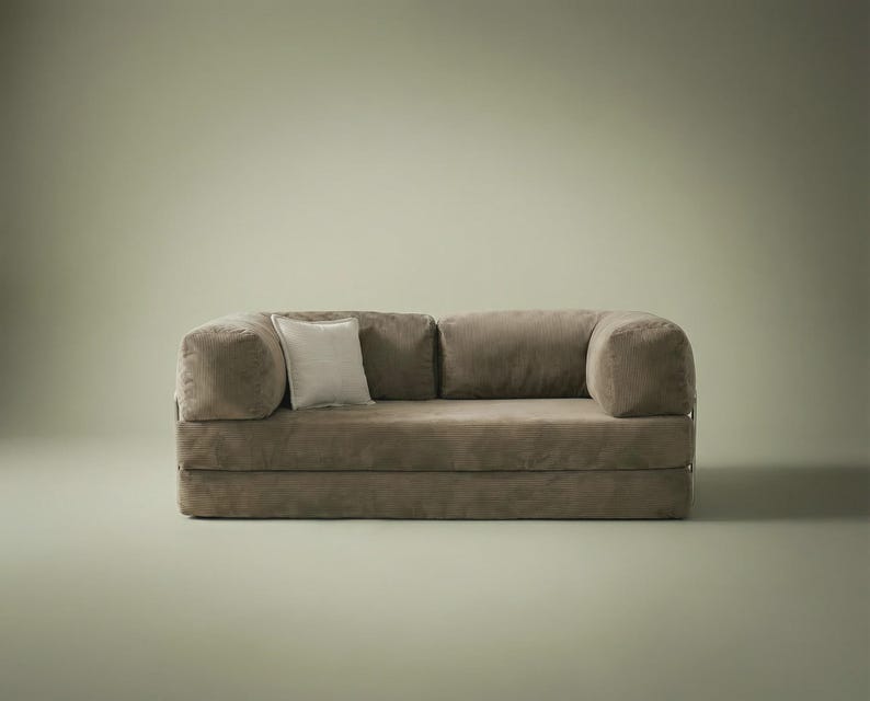 May include: A low-profile, taupe-colored sofa featuring a soft, textured fabric finish, rounded armrests, and two back cushions. A single, smaller square throw pillow in a lighter, off-white shade is positioned against the left back cushion.