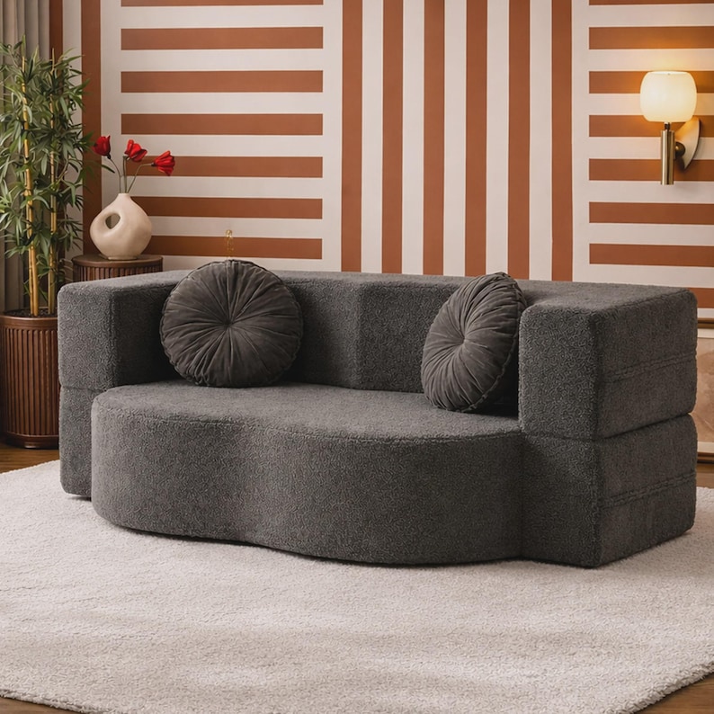 May include: A charcoal gray sofa upholstered in a textured, fuzzy fabric. The sofa features a curved, kidney-bean-shaped seat and two matching round, pleated throw pillows placed against the backrest. The design includes blocky armrests.