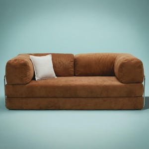 May include: A brown corduroy sofa featuring a low-profile design with thick, rounded armrests and a chrome metal frame. A single square white throw pillow with a subtle grid pattern rests against the left side of the back cushion.