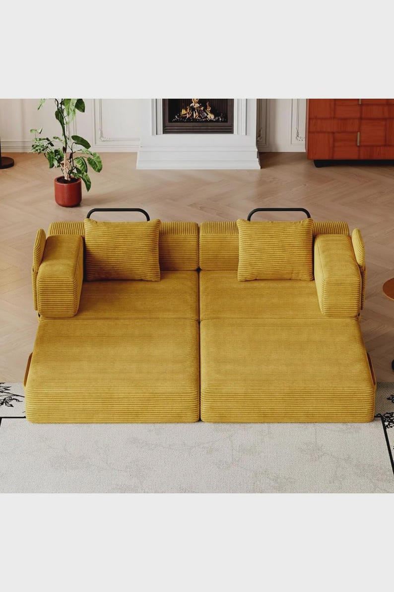 May include: A mustard yellow corduroy sofa bed featuring a low-profile design with two square cushions, two rectangular armrests, and two black metal handles visible at the back. The upholstery has a distinct horizontal ribbed texture.