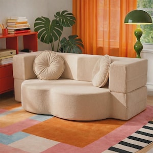 May include: A beige, curved, low-profile sofa upholstered in a textured fabric. The sofa features a rounded, cloud-like seat base and two matching round, tufted throw pillows placed against the backrest, providing a soft and neutral seating option.