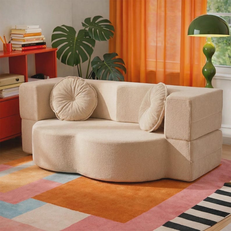 May include: A beige, curved, low-profile sofa upholstered in a textured fabric. The sofa features a rounded, cloud-like seat base and two matching round, tufted throw pillows placed against the backrest, providing a soft and neutral seating option.