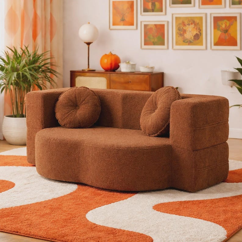 May include: A brown, textured, boucle-style loveseat with a curved, low-profile back and seat. The sofa features two matching round, tufted throw pillows placed against the backrest, providing a soft, uniform appearance in a solid earth tone.