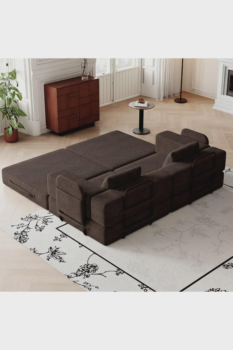 May include: A dark brown modular sofa bed with a textured corduroy fabric finish. The piece features a low-profile design with multiple square back cushions and a wide, flat base that extends outward to create a large, level sleeping surface.