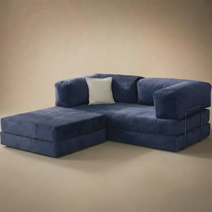 May include: A deep blue modular sofa featuring a low-profile design with thick, plush cushions and a matching chaise lounge section. A single square off-white throw pillow is placed in the corner. The frame includes a thin, dark metal support.