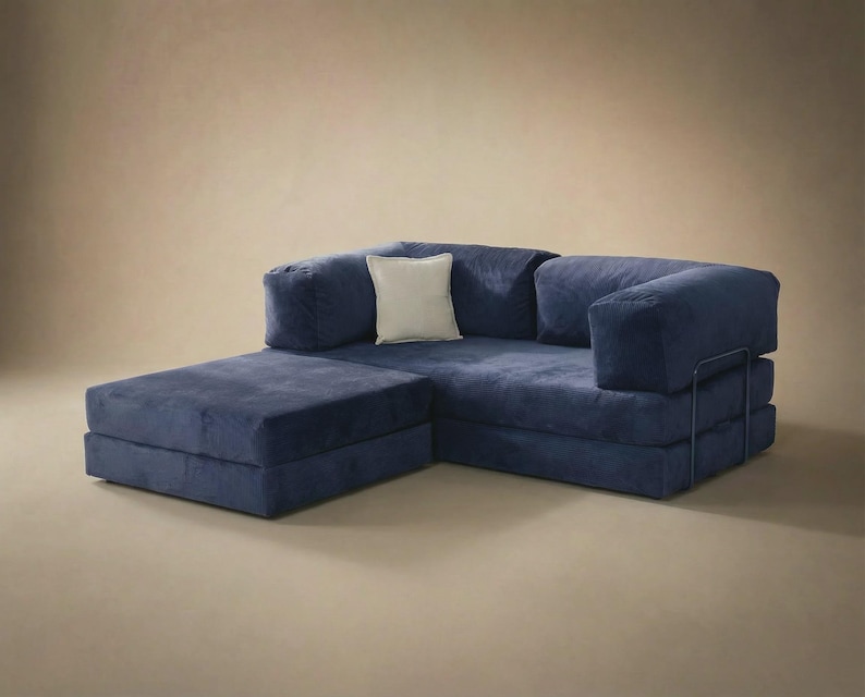 May include: A deep blue modular sofa featuring a low-profile design with thick, plush cushions and a matching chaise lounge section. A single square off-white throw pillow is placed in the corner. The frame includes a thin, dark metal support.