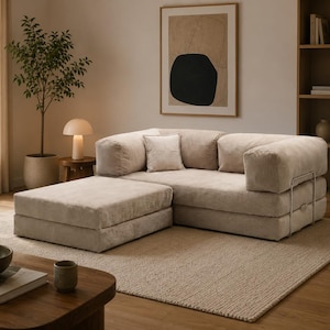 May include: A modular beige sofa with a matching ottoman, featuring a low-profile design, thick cushioned seating, and a sturdy metal frame structure. The upholstery has a soft, textured finish, and the set includes a single square throw pillow.