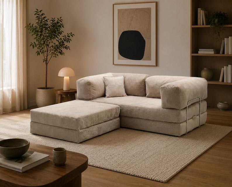 May include: A modular beige sofa with a matching ottoman, featuring a low-profile design, thick cushioned seating, and a sturdy metal frame structure. The upholstery has a soft, textured finish, and the set includes a single square throw pillow.