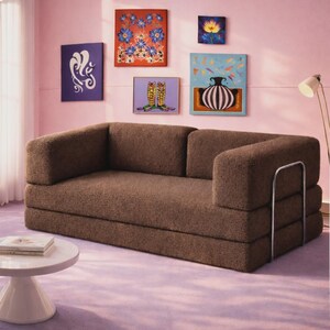 May include: A brown, textured, low-profile sofa with a rectangular backrest and seat, featuring a silver metal frame on the right side. The sofa is positioned in a room with pink walls and a light purple floor, with several framed art pieces on the wall.