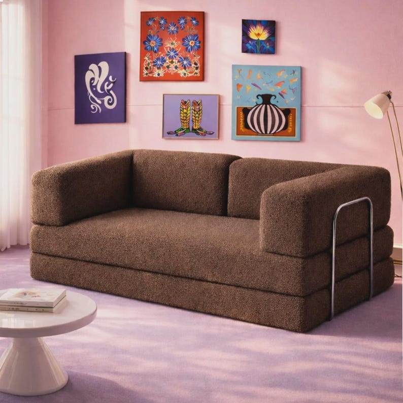 May include: A brown, textured, low-profile sofa with a rectangular backrest and seat, featuring a silver metal frame on the right side. The sofa is positioned in a room with pink walls and a light purple floor, with several framed art pieces on the wall.