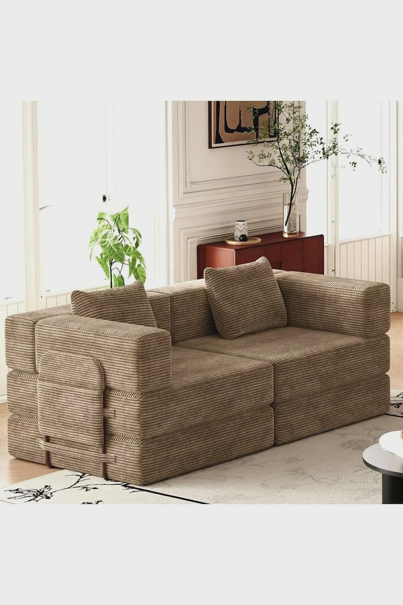 May include: A modular sofa upholstered in brown corduroy fabric, featuring a low-profile design with two square back cushions and a side panel attached with straps. The sofa consists of two distinct sections joined together to form a loveseat.