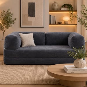 May include: A dark blue corduroy sofa with rounded armrests and a low-profile design. The sofa features a single square off-white throw pillow with a subtle grid pattern. The upholstery has a distinct vertical ribbed texture throughout the frame.