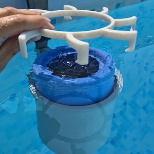 Könnte beinhalten: Ein weißer Pool-Skimmer-Aufsatz aus Kunststoff mit einem kreisförmigen Rahmen und nach außen gerichteten Zinken, der über einem blauen, zylindrischen Schwimmkörper positioniert ist, welcher teilweise im hellblauen Wasser schwimmt.