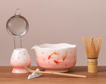 May include: A matcha tea set with a pink and white gradient bowl featuring koi fish illustrations. The set includes a bamboo whisk, a ceramic tea scoop, a stainless steel strainer, and a small ceramic holder for the scoop.