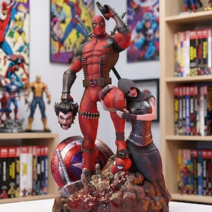 Puede incluir: Una estatua coleccionable detallada de Deadpool con traje rojo y negro sosteniendo una cabeza cortada, junto a una figura encapuchada de negro. La base incluye un escudo del Capitán América, un martillo y una máscara de Spider-Man sobre rocas.