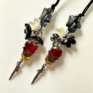 May include: A pair of dangling earrings on black cords, featuring faceted red and amber beads, dark grey star-shaped beads, and translucent flower-shaped accents. Each earring terminates in a silver-toned metal sword-shaped charm.