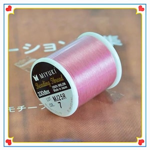 May include: A spool of pink Miyuki beading thread made of 100% nylon. The label on the white plastic spool indicates 330dtex, lot MJ25R, and color 7. The thread is wound tightly around the spool, displaying a smooth, shiny, and vibrant pink texture.