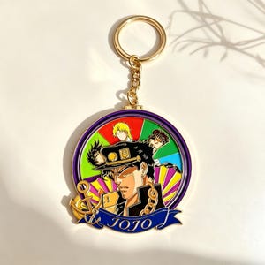 Può includere: Un portachiavi circolare in metallo dorato con un'illustrazione colorata in smalto di personaggi anime. La figura centrale indossa un berretto e un cappotto scuri con catene dorate. Un nastro blu in basso mostra il testo JOJO in bianco.