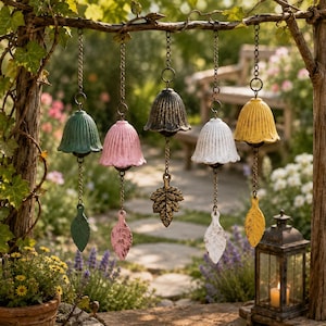 May include: A selection of colourful wind chimes, with bell-shaped tops and leaf pendants, hanging from a branch. The chimes are in shades of green, pink, black, white, and yellow. They are suspended by chains and displayed in a garden, creating a decorative outdoor feature.