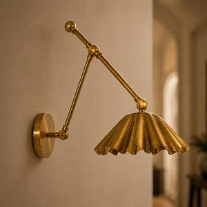 Puede incluir: Una lámpara de pared de latón con un brazo articulado ajustable con múltiples puntos de pivote y una pantalla ancha, plisada y en forma de campana. El aplique tiene una placa de pared circular y un acabado metálico.