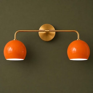 May include: A wall-mounted light fixture featuring a horizontal brass-toned metal bar and a circular mounting plate. Two bright orange, dome-shaped glass shades are attached to the ends of the bar, each pointing downward to emit light.