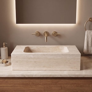 May include: A rectangular beige travertine stone vessel sink with a natural porous texture. The sink is mounted on a countertop and paired with a wall-mounted brushed gold faucet featuring two side handles and a central curved spout.