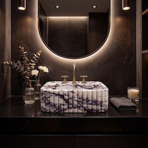 Könnte beinhalten: Rechteckiges Waschbecken aus weißem Marmor mit violetten Adern und geriffelter Oberfläche. Goldfarbene Armaturen und ein runder Spiegel mit Hintergrundbeleuchtung. Ein luxuriöses Badezimmer-Design.
