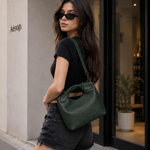 Könnte beinhalten: Eine dunkelwaldgrüne Handtasche mit einer strukturierten, lederartigen Oberfläche, die einen in den Körper der Tasche integrierten, strukturierten Tragegriff sowie einen abnehmbaren Schulterriemen mit Metallclips an den Seiten aufweist.