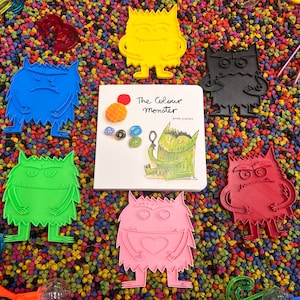 May include: A selection of brightly coloured monster-shaped cutouts in blue, yellow, black, red, green, and pink, surrounding a book titled "The Colour Monster." The background is filled with small, multicoloured beads, with various tools scattered around.