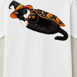 Op de afbeelding: Een wit t-shirt met een afbeelding van een slapende zwart-witte kat die een zwarte en oranje heksenhoed draagt en een bijpassende cape versierd met oranje pompoenen, gele sterren en halve manen.