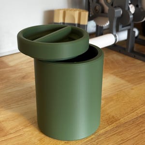 May include: An olive green cylindrical container with a matching lid. The lid is round and divided, slightly open to show the inside. It rests on a wooden surface. Other objects are visible in the background.