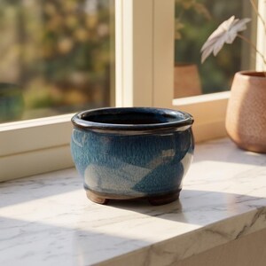 May include: A round ceramic planter with a glossy finish featuring a deep blue and teal gradient glaze. The bowl-shaped vessel has a dark brown rim and sits on three small, integrated feet, showcasing a smooth, reflective surface texture.