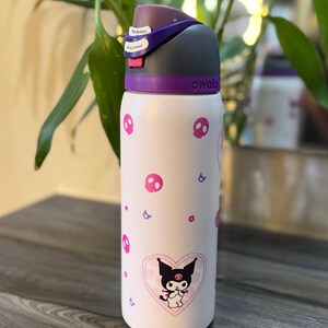 Puede incluir: Una botella de agua Owala blanca con un gr&aacute;fico del personaje Kuromi dentro de un coraz&oacute;n rosa, rodeado de peque&ntilde;os iconos de calaveras y flores rosas. La botella tiene una tapa gris oscuro y morada con bot&oacute;n y cierre de seguridad.