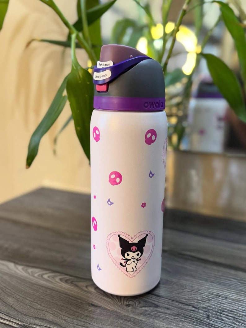 Puede incluir: Una botella de agua Owala blanca con un gr&aacute;fico del personaje Kuromi dentro de un coraz&oacute;n rosa, rodeado de peque&ntilde;os iconos de calaveras y flores rosas. La botella tiene una tapa gris oscuro y morada con bot&oacute;n y cierre de seguridad.