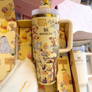 May include: A yellow Stanley tumbler featuring Winnie the Pooh characters and the text "Winnie the Pooh". The tumbler has a handle and a bee-shaped topper. The tumbler is packaged in a themed box.