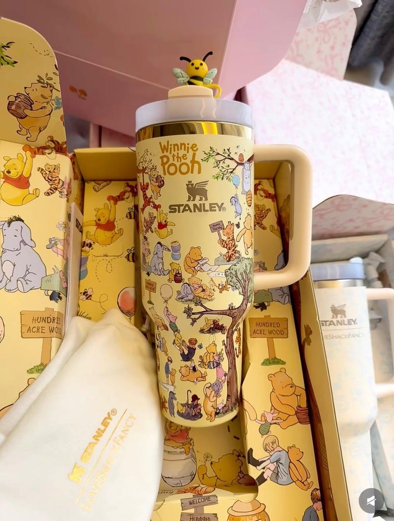 May include: A yellow Stanley tumbler featuring Winnie the Pooh characters and the text "Winnie the Pooh". The tumbler has a handle and a bee-shaped topper. The tumbler is packaged in a themed box.