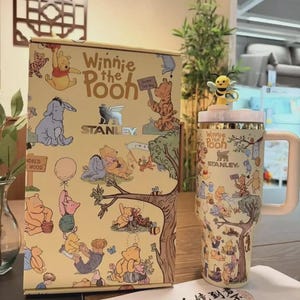 May include: A Winnie the Pooh themed Stanley tumbler and matching box. The tumbler has a white handle and a yellow lid with a bee figurine. The box and tumbler feature illustrations of Winnie the Pooh and friends.