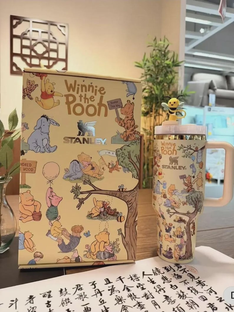 May include: A Winnie the Pooh themed Stanley tumbler and matching box. The tumbler has a white handle and a yellow lid with a bee figurine. The box and tumbler feature illustrations of Winnie the Pooh and friends.